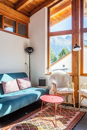 A cozy living room with a blue sofa, a red table, and a view of mountains through large windows. - La Cabane Verte – Sunny family apartment near St. Moritz & Ski Slopes (Celerina/Schlarigna)