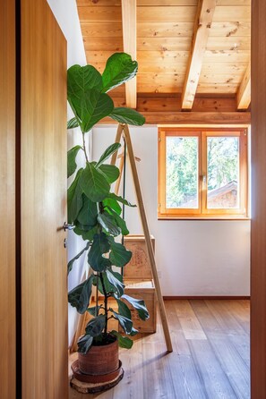 A room with wooden flooring, a large window, and a potted plant on a wooden stand. - La Cabane Verte – Sunny family apartment near St. Moritz & Ski Slopes (Celerina/Schlarigna)