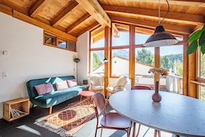 Et hyggeligt rum med træloft, sofa, spisebord og store vinduer med udsigt til et bjerglandskab.
