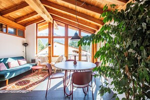 A room with a wooden ceiling, large windows, a round table, and chairs. - The Green Hut – Sunny Family Apartment Near St. Moritz & the Ski Slopes (Celerina/Schlarigna)