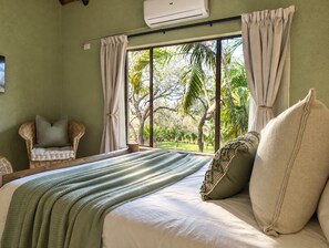 A bedroom with a large window, a bed with a green blanket, a wicker chair, and a view of greenery outside.