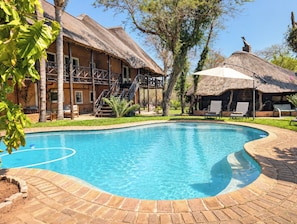 A swimming pool with a brick border, a wooden deck, and a thatched-roof building with a staircase. - Leriti Safari House (Marloth Park)