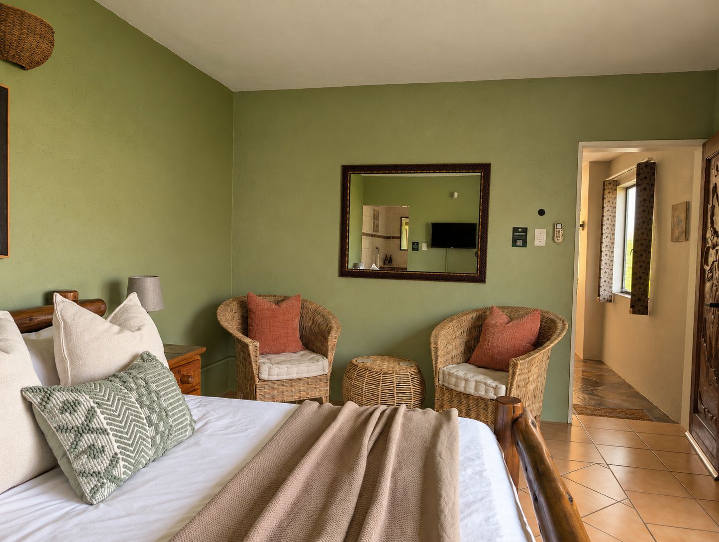 A bedroom with a bed, two wicker chairs, a mirror, and a television.