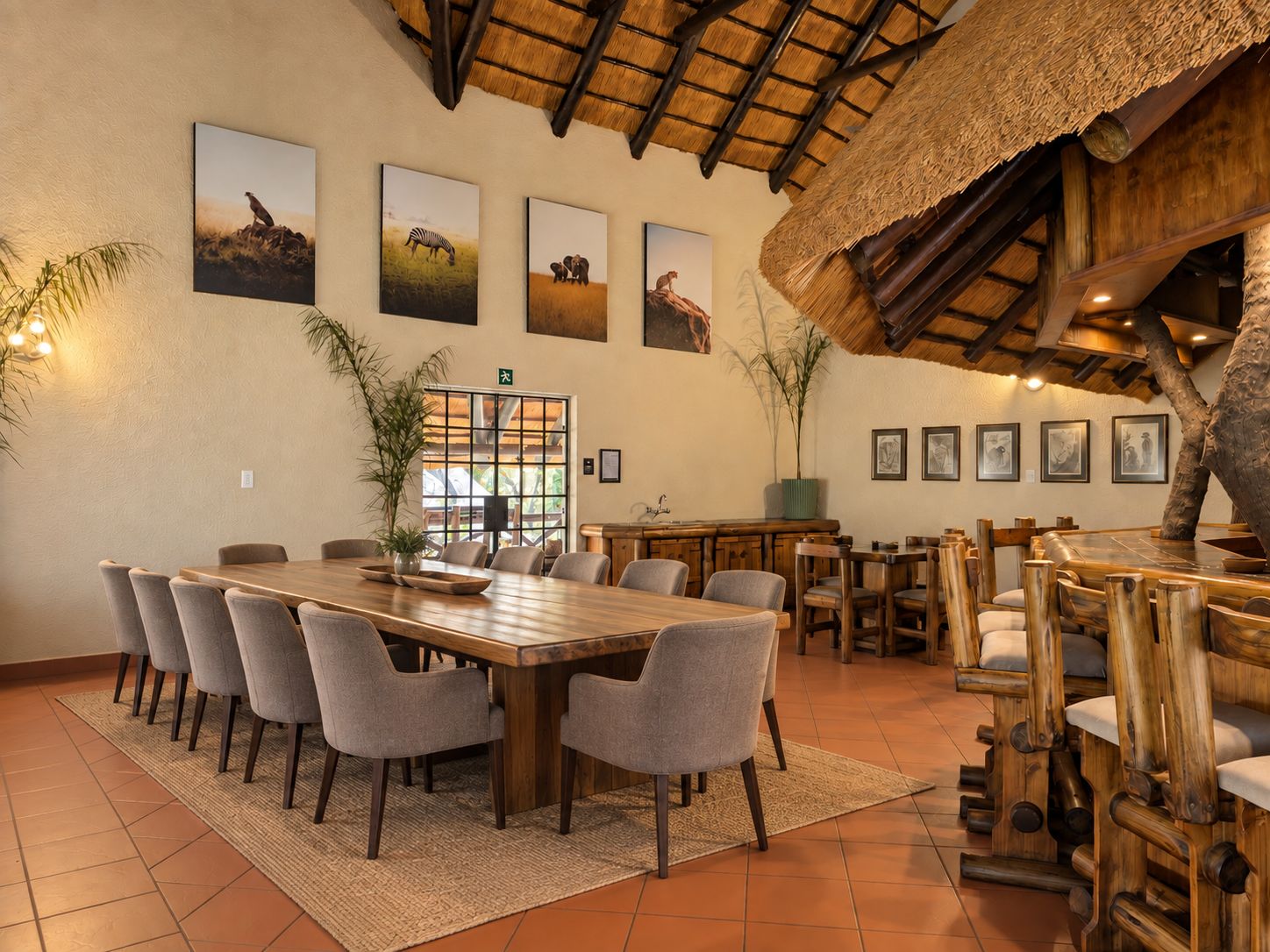 A dining area with a large wooden table surrounded by chairs, framed wildlife pictures on the wall, and a thatched roof structure.