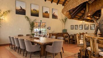 A dining area with a large wooden table surrounded by chairs, framed wildlife pictures on the wall, and a thatched roof structure.