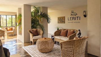 Uma sala de estar ampla com móveis de vime, uma grande mesa de centro trançada e uma placa na parede com a inscrição 'LERITI SAFARI HOUSE'.