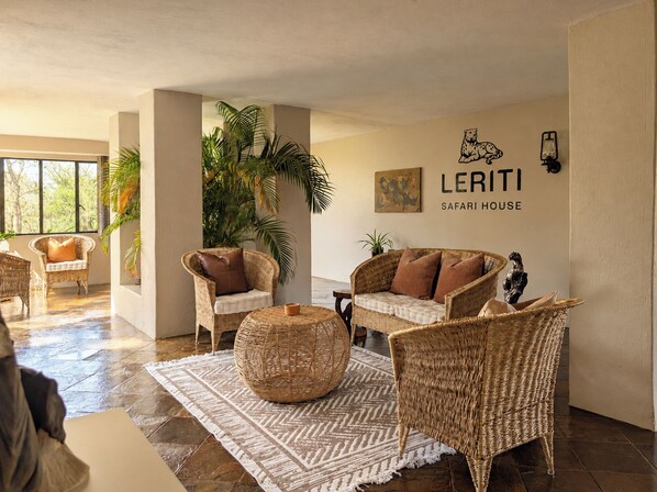 Un vaste espace de vie agrémenté de meubles en osier, d’une grande table basse en fibres tressées et d’une enseigne fixée au mur portant l’inscription « LERITI SAFARI HOUSE ».