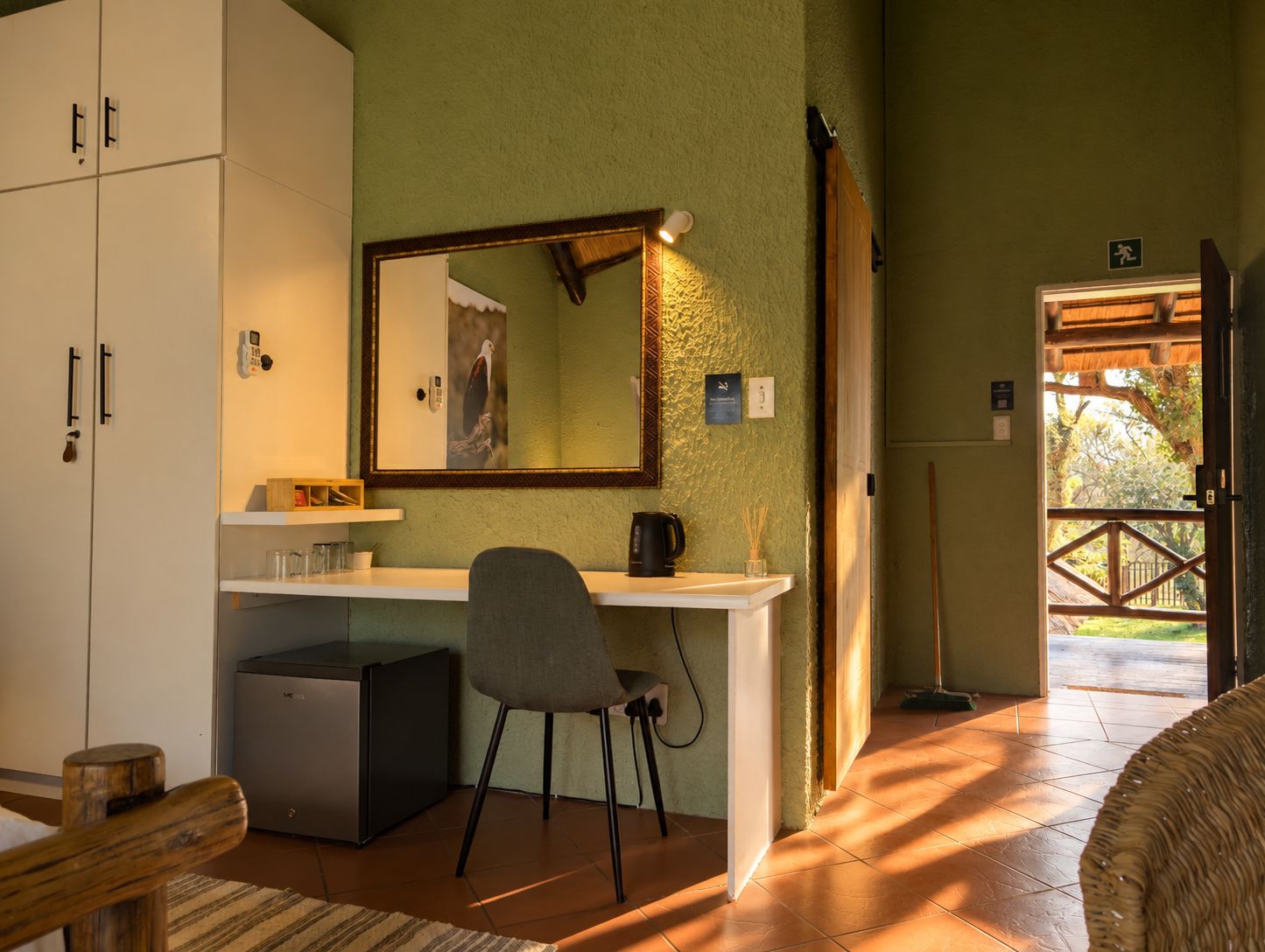 Quarto com escrivaninha, cadeira, armários e espelho. Uma porta dá para o exterior.