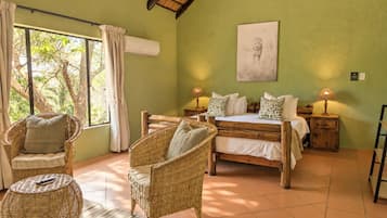 Quarto com cama, duas cadeiras de vime, janela com cortinas, ar-condicionado fixado na parede e um quadro emoldurado.
