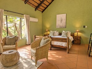 Une pièce comprenant un lit, deux fauteuils en osier, une fenêtre avec des rideaux, un climatiseur fixé au mur et un tableau encadré.