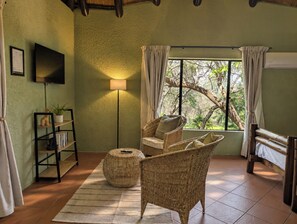 A room with a wicker chair, a floor lamp, a bookshelf, and a bed with a view of trees through a window.