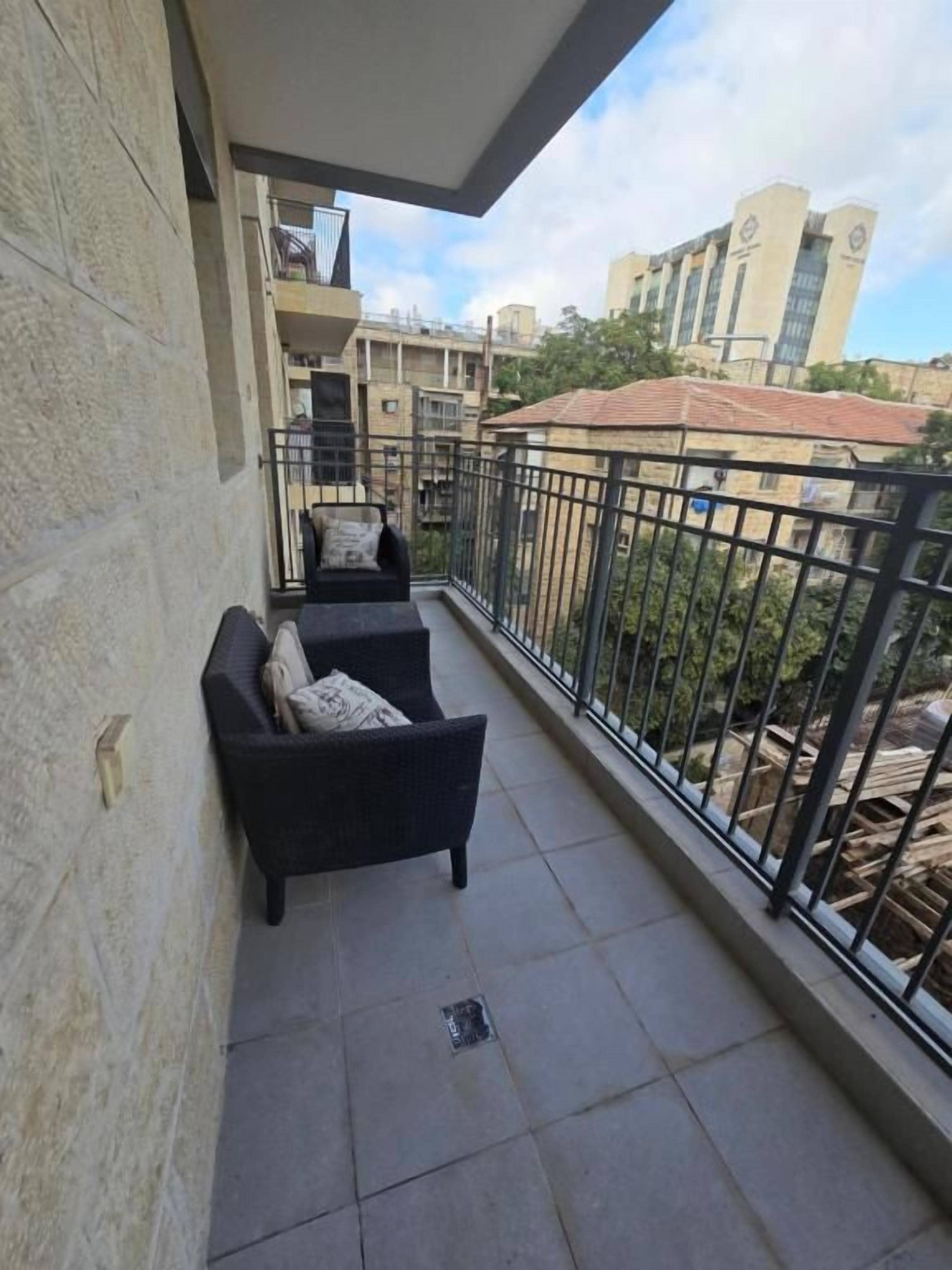 A balcony with a black wicker sofa and a small table, a metal railing, and a view of buildings in the background.