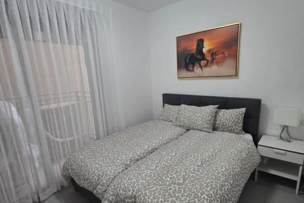 A bedroom with a bed, a nightstand, a painting on the wall, and a window with curtains.