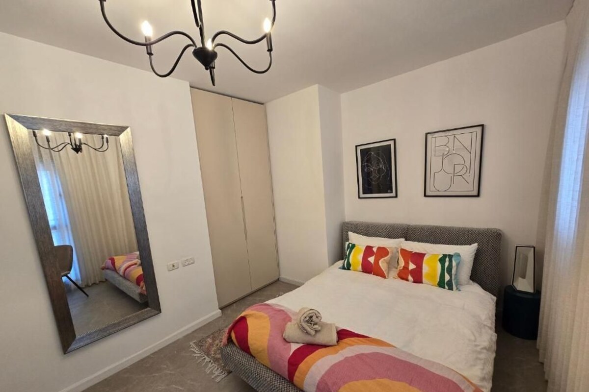 A bedroom with a bed, a large mirror, a chandelier, and two framed pictures on the wall.