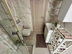A bathroom with a glass shower enclosure, a toilet, a sink, and a shelf with towels. - Belani Bay Apartment 11 | Balcony and Parking in Tivat (Tivat)