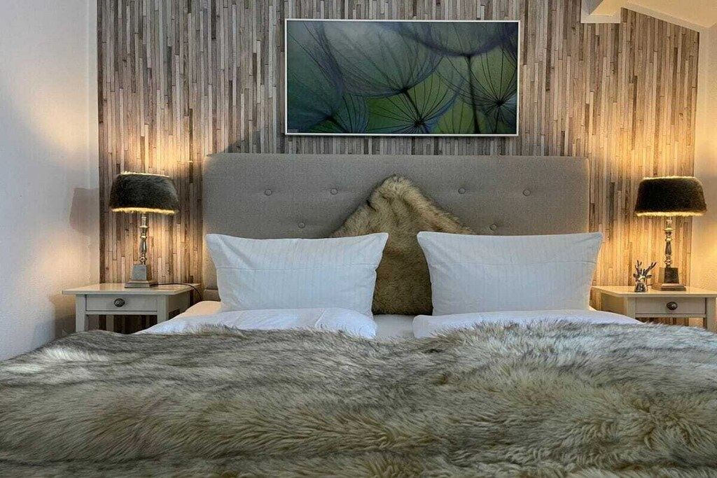 Quarto com cabeceira estofada, uma pintura abstrata grande, duas mesas de cabeceira com luminárias e um cobertor macio.