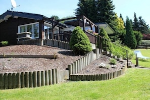 A two-story house with a deck, a satellite dish, and a landscaped garden with a stone retaining wall. - Holiday home with 2 bedrooms (Winterberg-Niedersfeld)