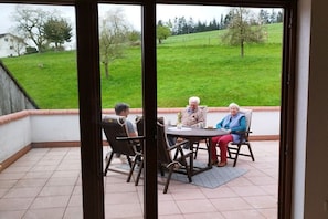 A patio with a table and chairs, a view of a green field, and a building in the background. - Holiday apartment with 3 bedrooms (Oberzent Airlenbach)
