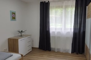 A bedroom with a bed, a wooden dresser, a nightstand, and a window with sheer curtains. - Holiday apartment with 3 bedrooms (Oberzent Airlenbach)