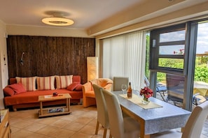 A living room with a sofa, a table, and a chair. There is a window with curtains and a view of greenery outside. - "Chargund": Holiday apartment "Chardonnay" (Herxheim)