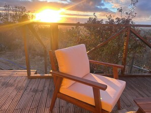 Um deck de madeira com uma única cadeira branca acolchoada, com vista para uma paisagem natural com árvores e um pôr do sol.