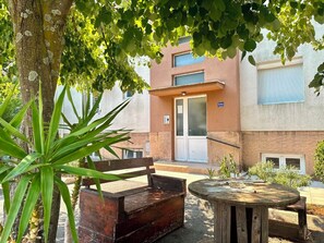 Un espace extérieur aménagé avec un banc et une table en bois, entouré de verdure et bordé par un bâtiment doté d’une porte et de fenêtres.