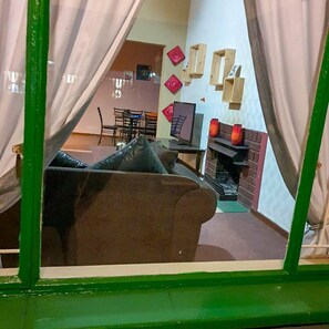 A room with a green-framed window, a brown sofa, a dining table with chairs, and a fireplace with red lanterns.