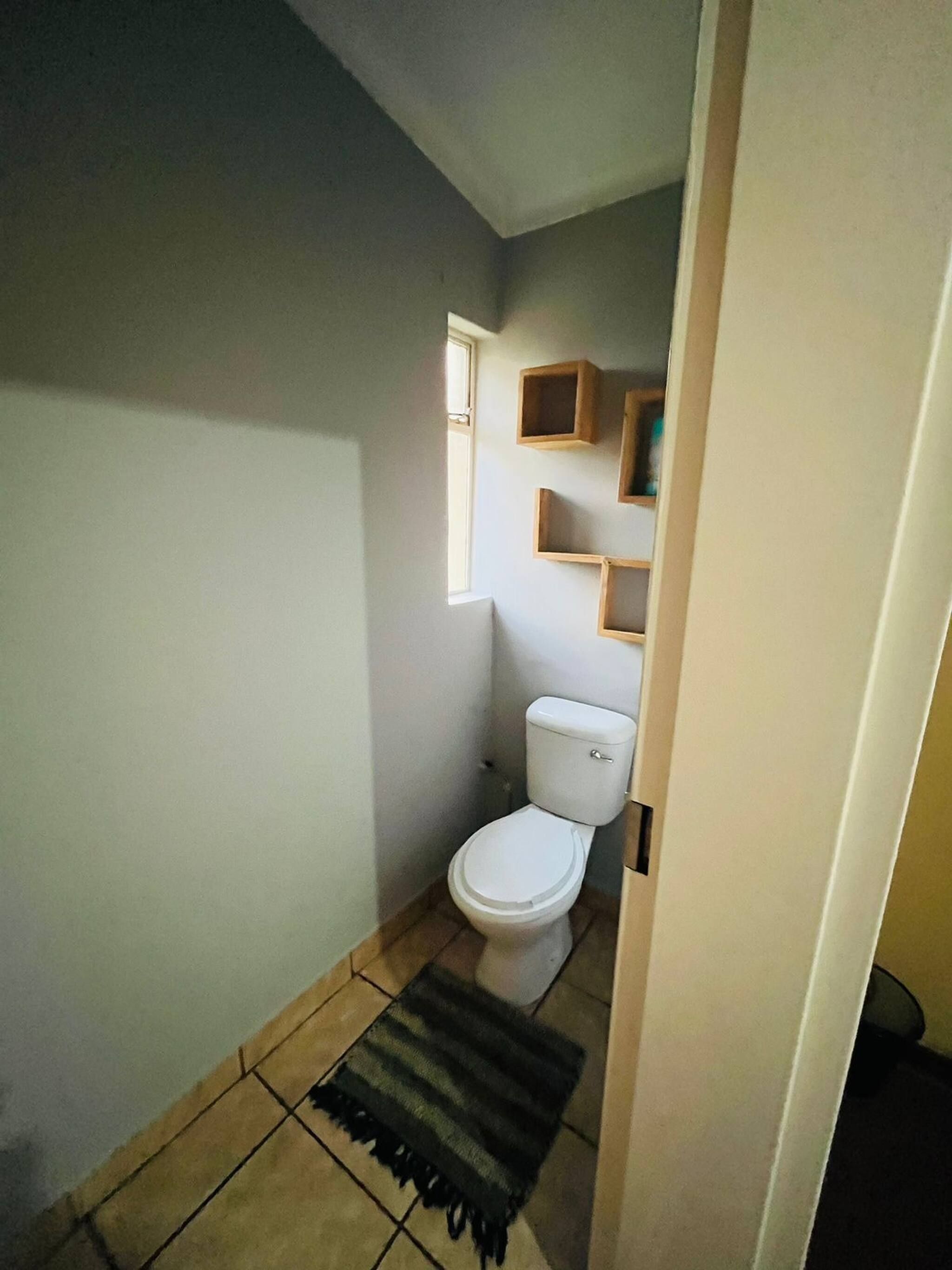 A small, narrow bathroom with a toilet, a window, and a shelf with a mirror.