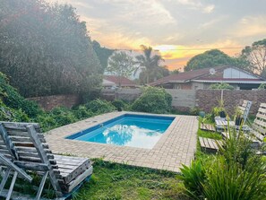 A rectangular swimming pool with a tiled edge, surrounded by a brick patio. There are wooden lounge chairs and a wooden deck with a ladder.