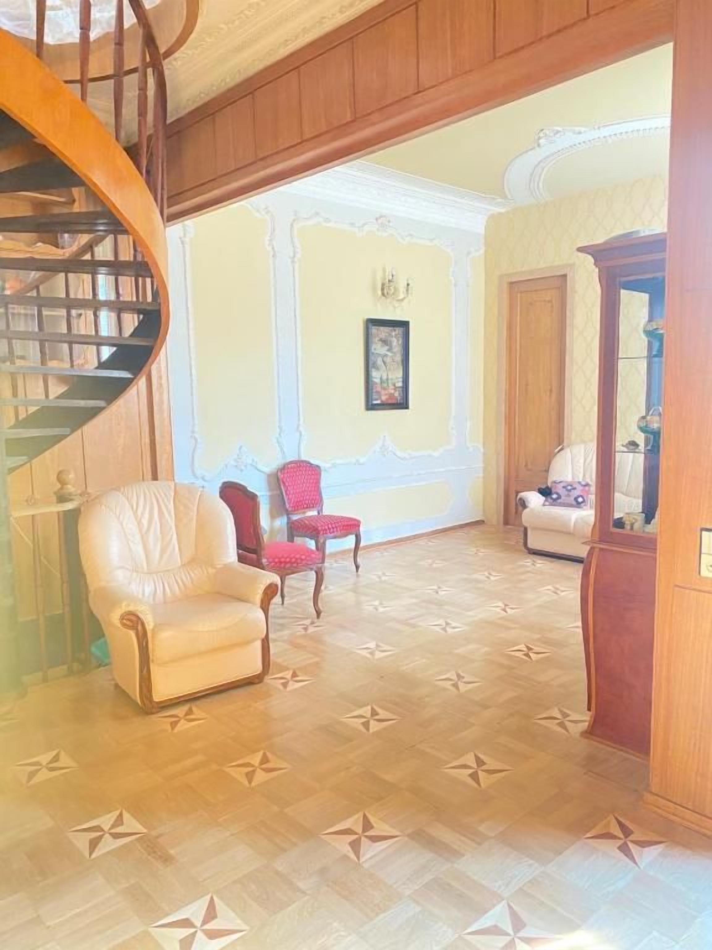 Una habitación con una escalera de caracol, un sillón blanco, una silla roja, un sofá y un aparador de madera.