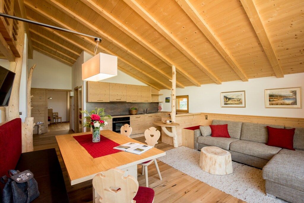 Ein gemütliches Interieur mit Holzbalkendecke, einem Essbereich mit roter Tischdecke, einem grauen Sofa mit roten Kissen und einer Küche im Hintergrund.
