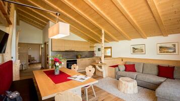 Ein gemütliches Interieur mit Holzbalkendecke, einem Essbereich mit roter Tischdecke, einem grauen Sofa mit roten Kissen und einer Küche im Hintergrund.
