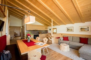 A cozy interior with a wooden ceiling, a dining area with a red table runner, a grey sofa with red cushions, and a kitchen in the background. - Apartment in Lajen with swimming pool (Lajen)
