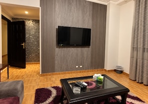 A modern living room with a flat-screen TV mounted on a textured wall, a glass coffee table with remote controls, and a plush rug. - Luxury Apartment in New Cairo (Cairo Governorate)