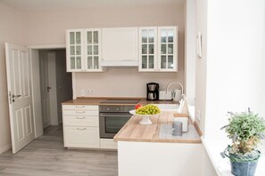 Een kleine keuken met witte kastjes, een inbouwoven, een koffiezetapparaat en een fruitschaal op het aanrecht.