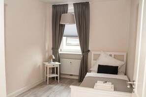 Ein Schlafzimmer mit Bett, Nachttisch, Lampe und Fenster mit Vorhängen.