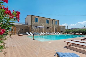 Un área junto a la piscina con sillones y sombrillas, un edificio de piedra con ventanas, y un cielo azul despejado.