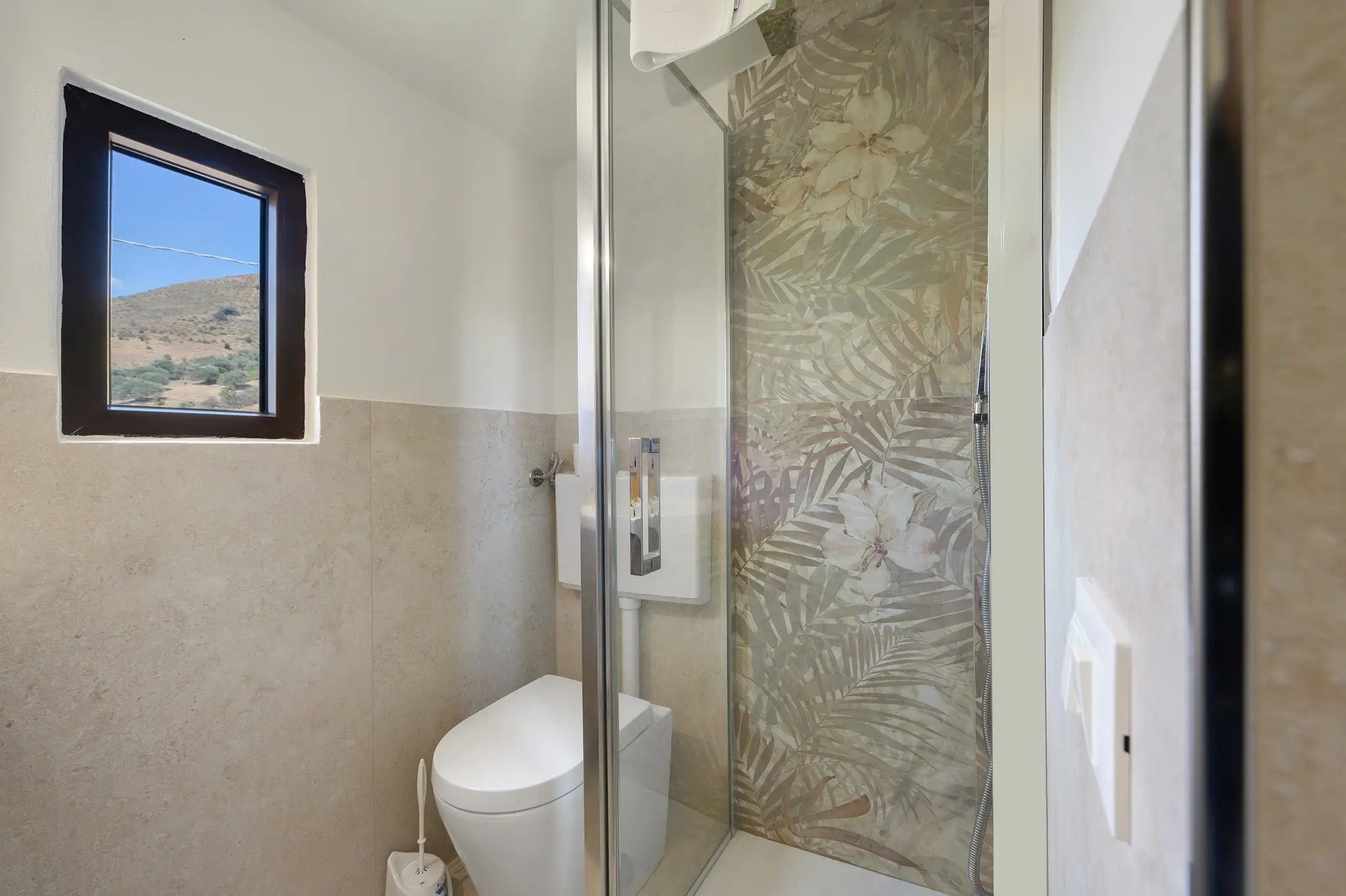 A small bathroom with a toilet, a shower with a glass door, and a window with a view of a desert landscape.