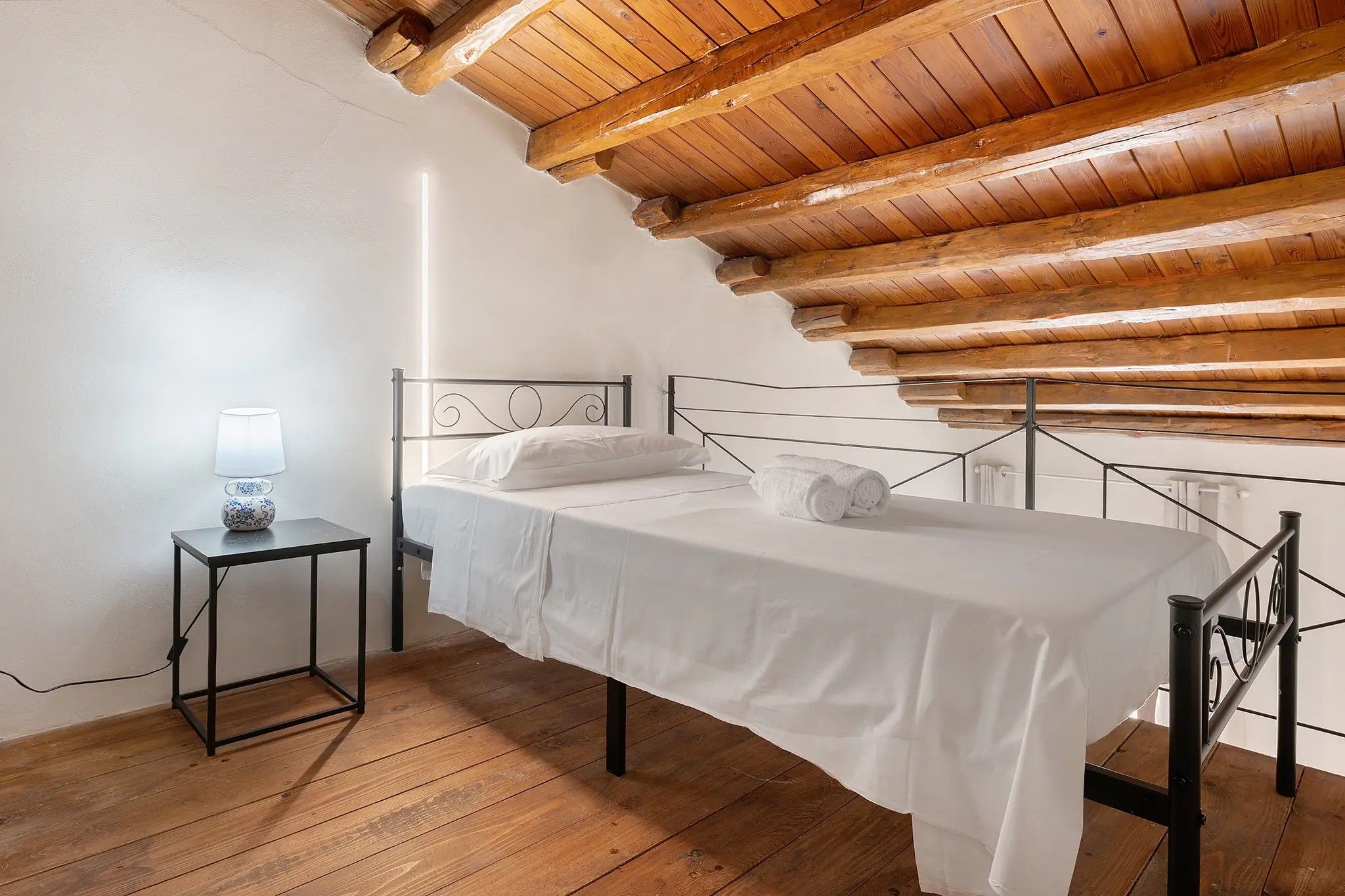 A single bed with a metal frame, a bedside table with a lamp, and a wooden ceiling.