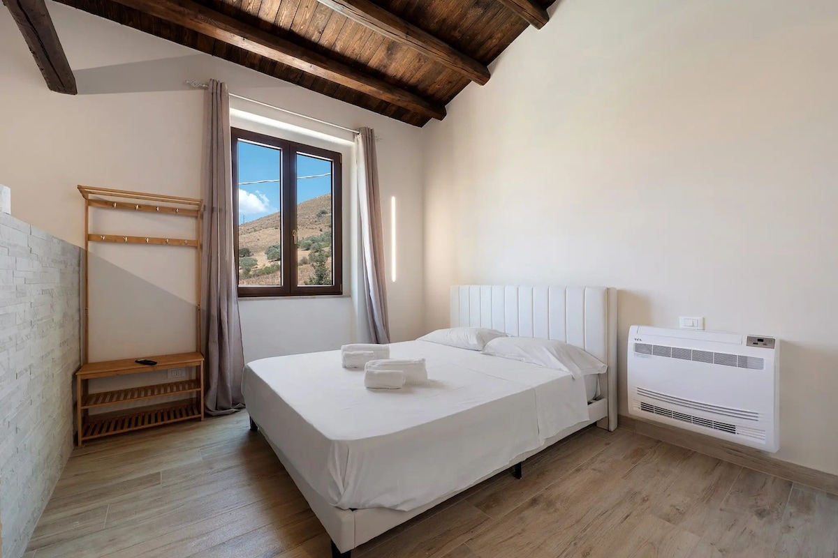A bedroom with a bed, a wooden bench, a window with curtains, and an air conditioning unit.