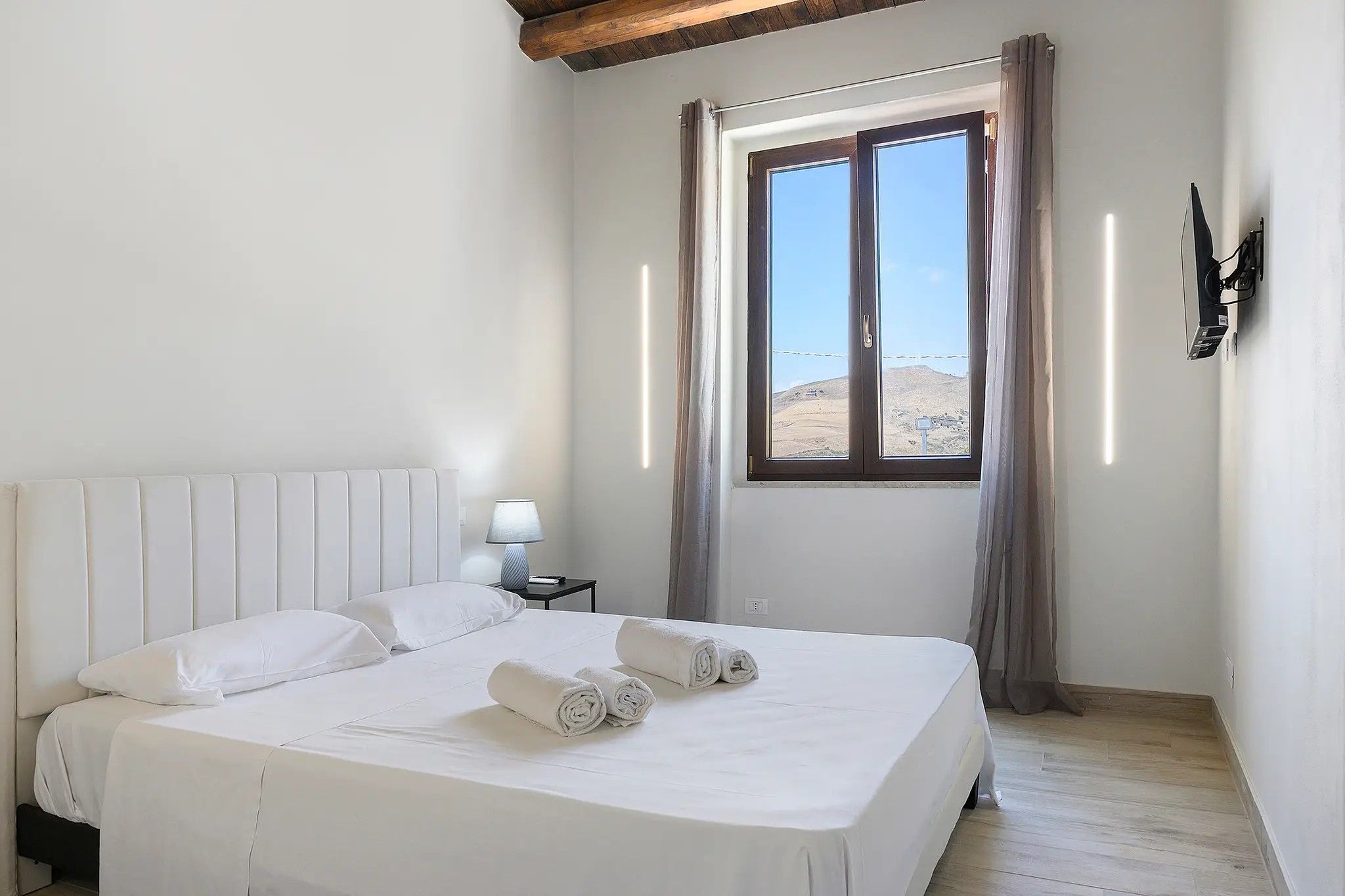 Ein Hotelzimmer mit einem großen Bett, einem Nachttisch, einer Lampe, einem Fenster mit Vorhängen und Blick auf eine Landschaft.