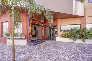 Entrada de un hotel con un patio empedrado, cerca viva blanca y una palmera.