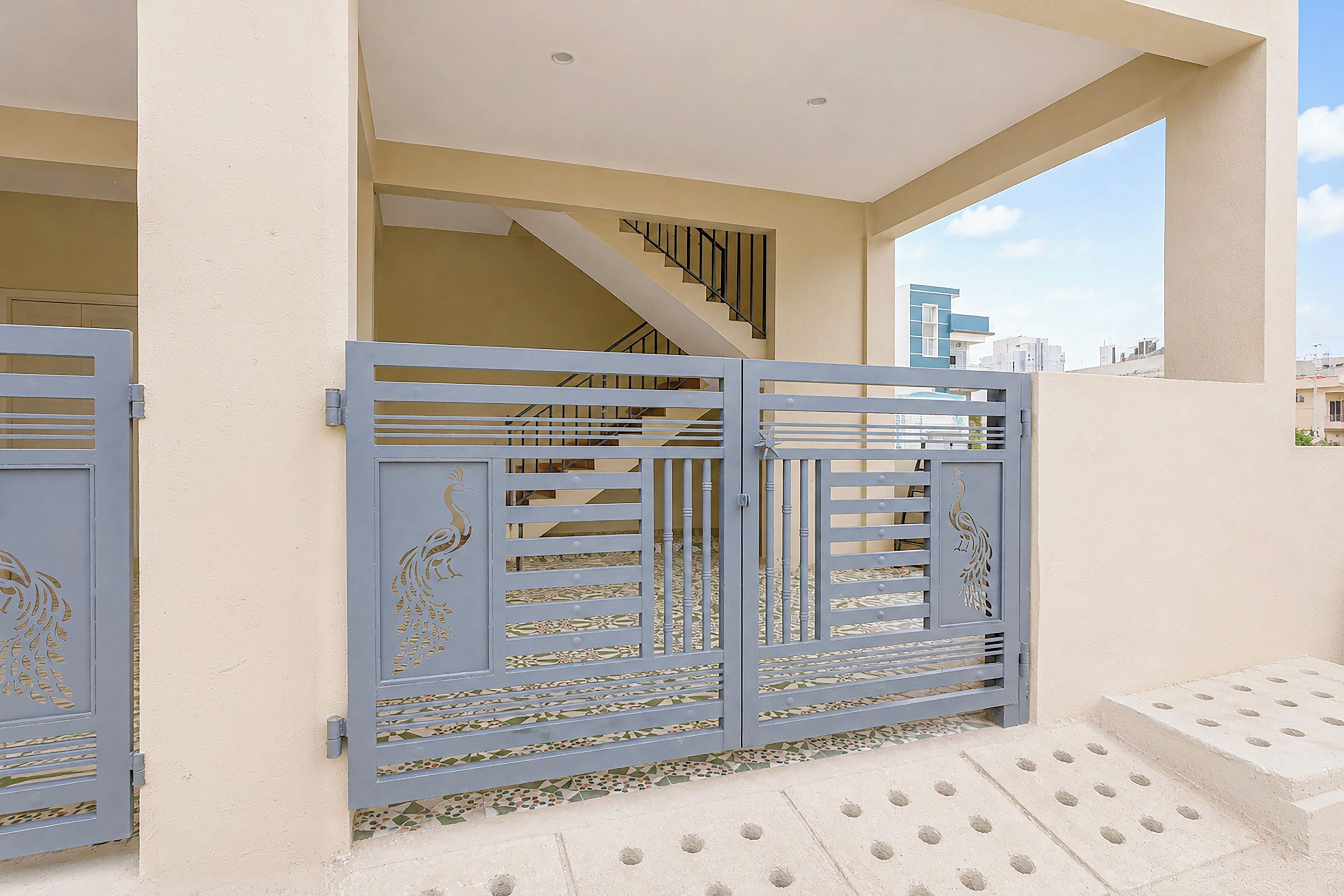 Una entrada protegida con ornamentación metálica, una escalera que sube a un nivel superior y una vista de un paisaje urbano a través de una ventana.