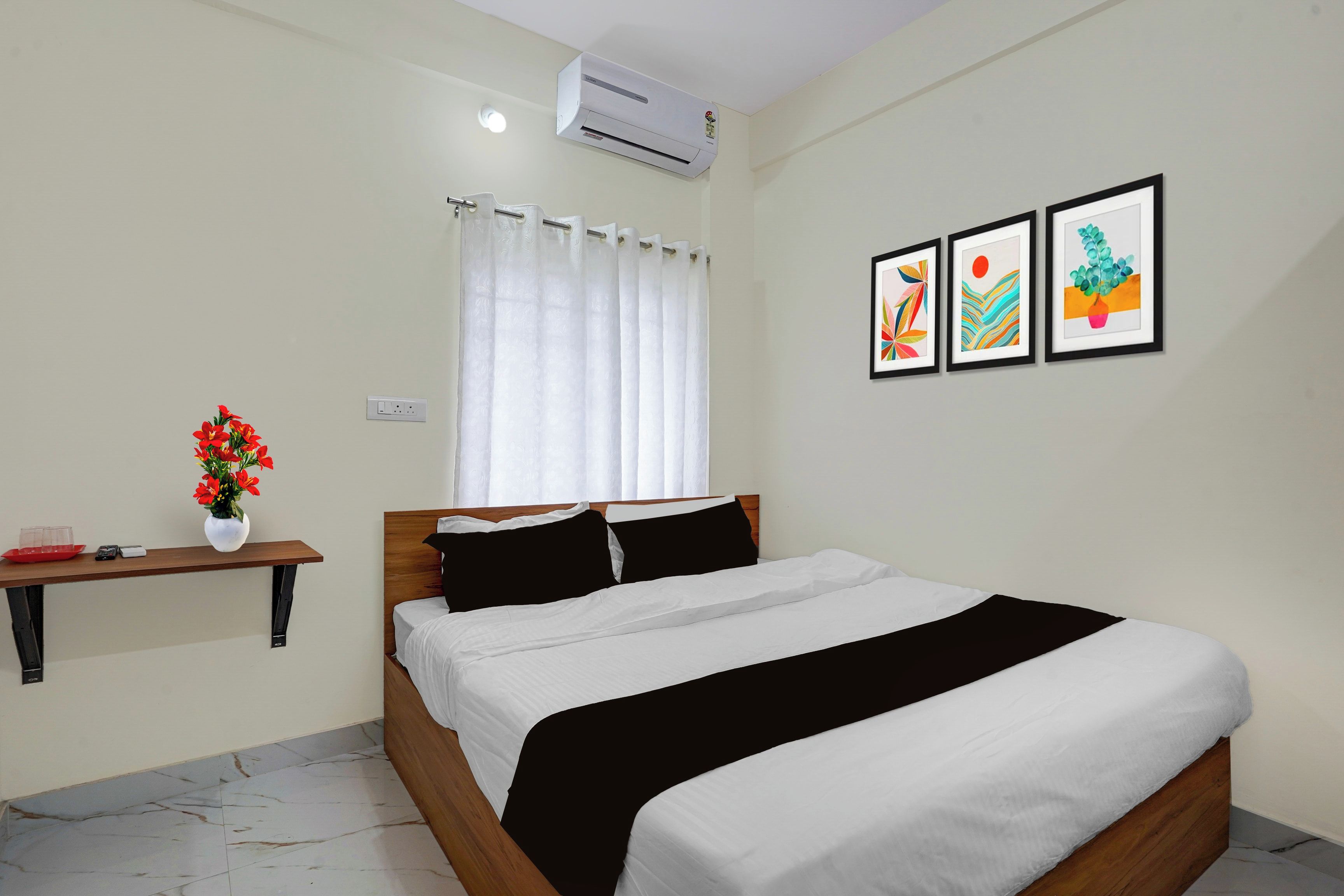 Um quarto com cama, mesa de cabeceira, janela com cortinas, ar condicionado e três quadros na parede.