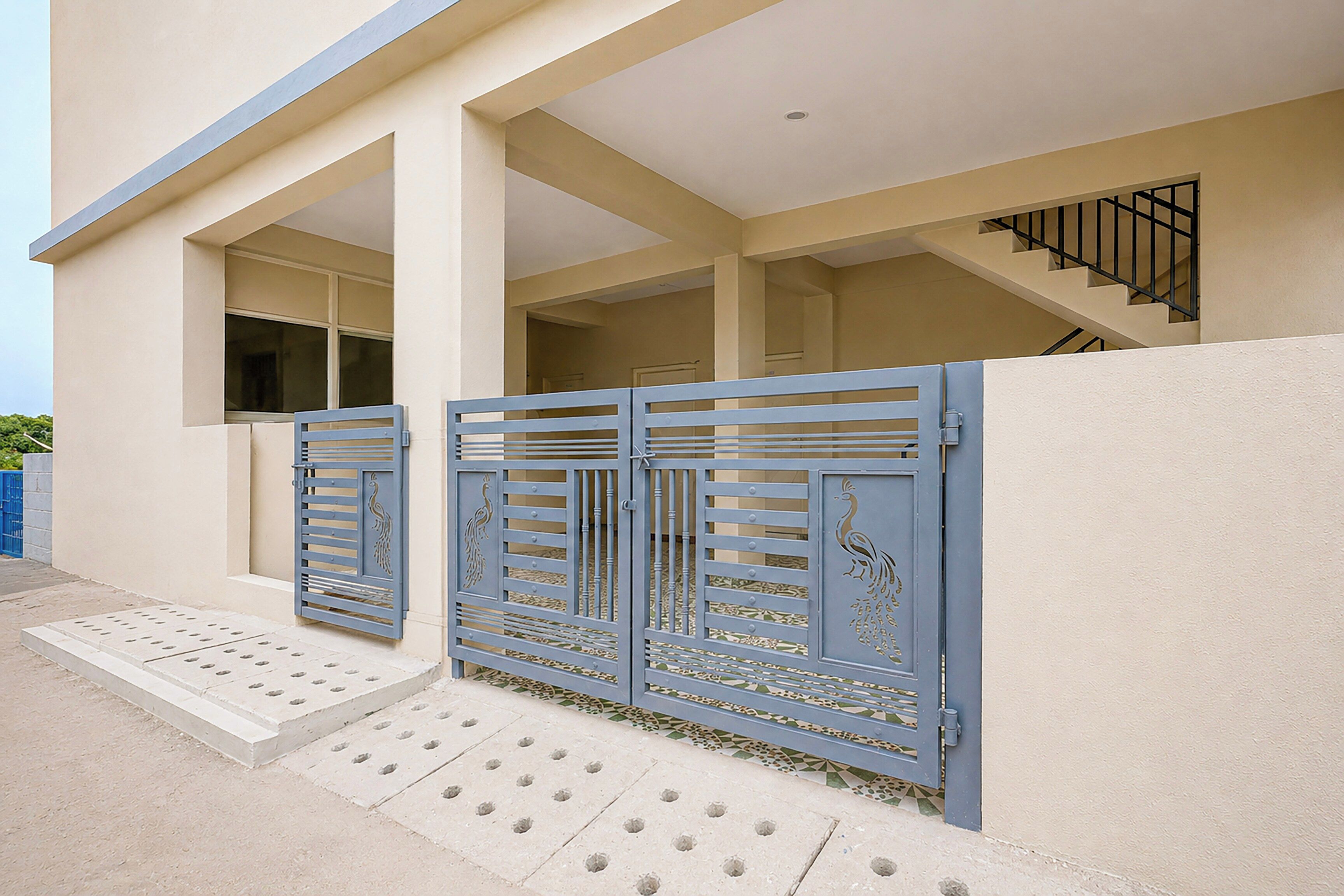 Un edificio moderno con fachada de color claro, una puerta azul con diseños de pavo real y un camino de hormigón que conduce a la entrada.