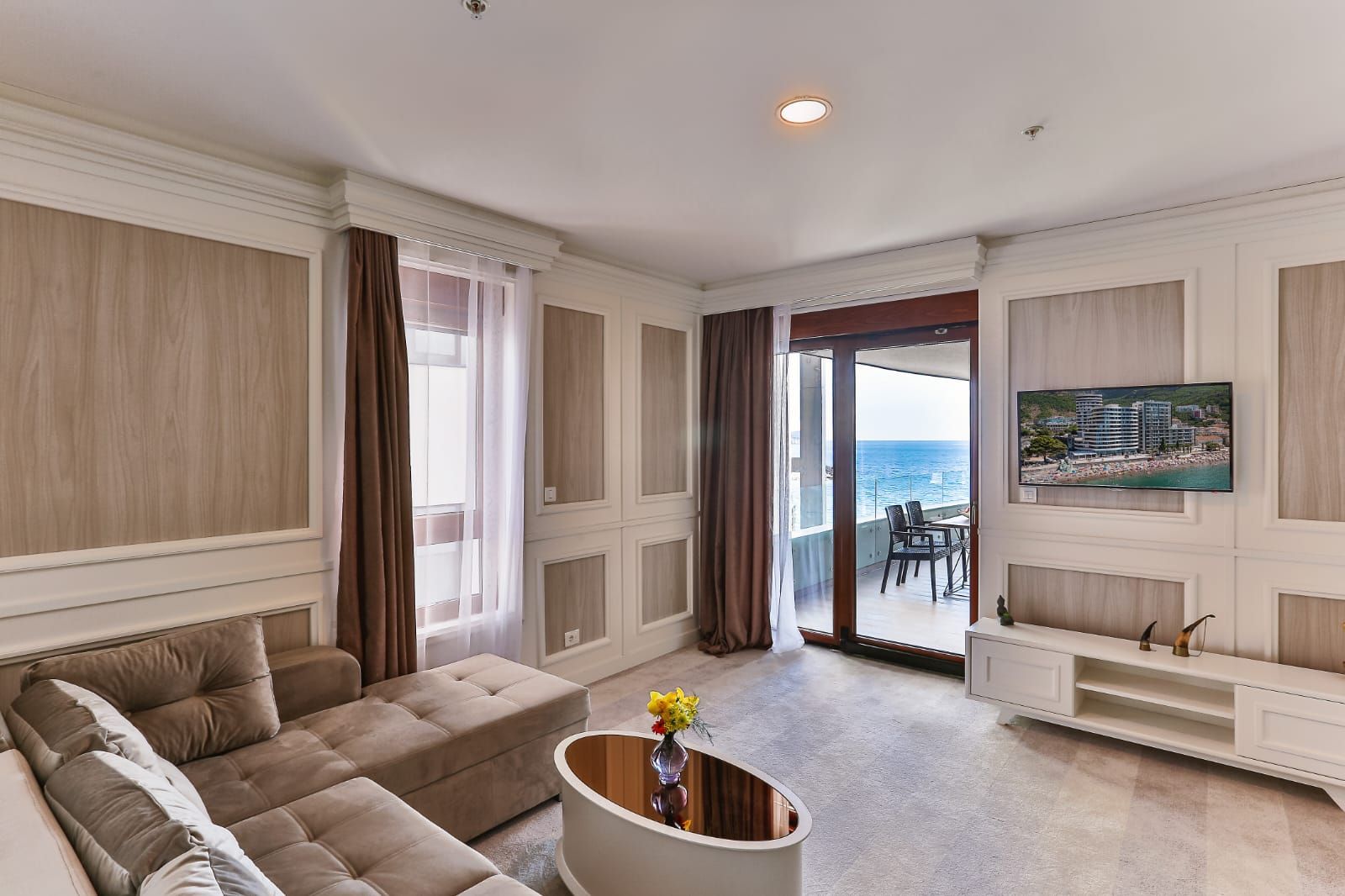 A spacious living room with a large sofa, a coffee table with flowers, a TV mounted on the wall, and a view of the sea through the glass door.