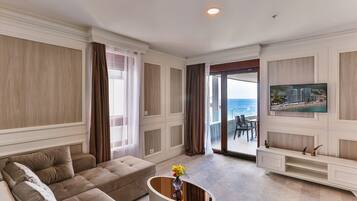 A spacious living room with a large sofa, a coffee table with flowers, a TV mounted on the wall, and a view of the sea through the glass door.