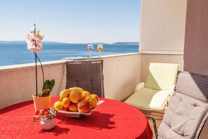 Een balkon met een rode tafel, een schaal fruit, een bloempot en twee glazen wijn.