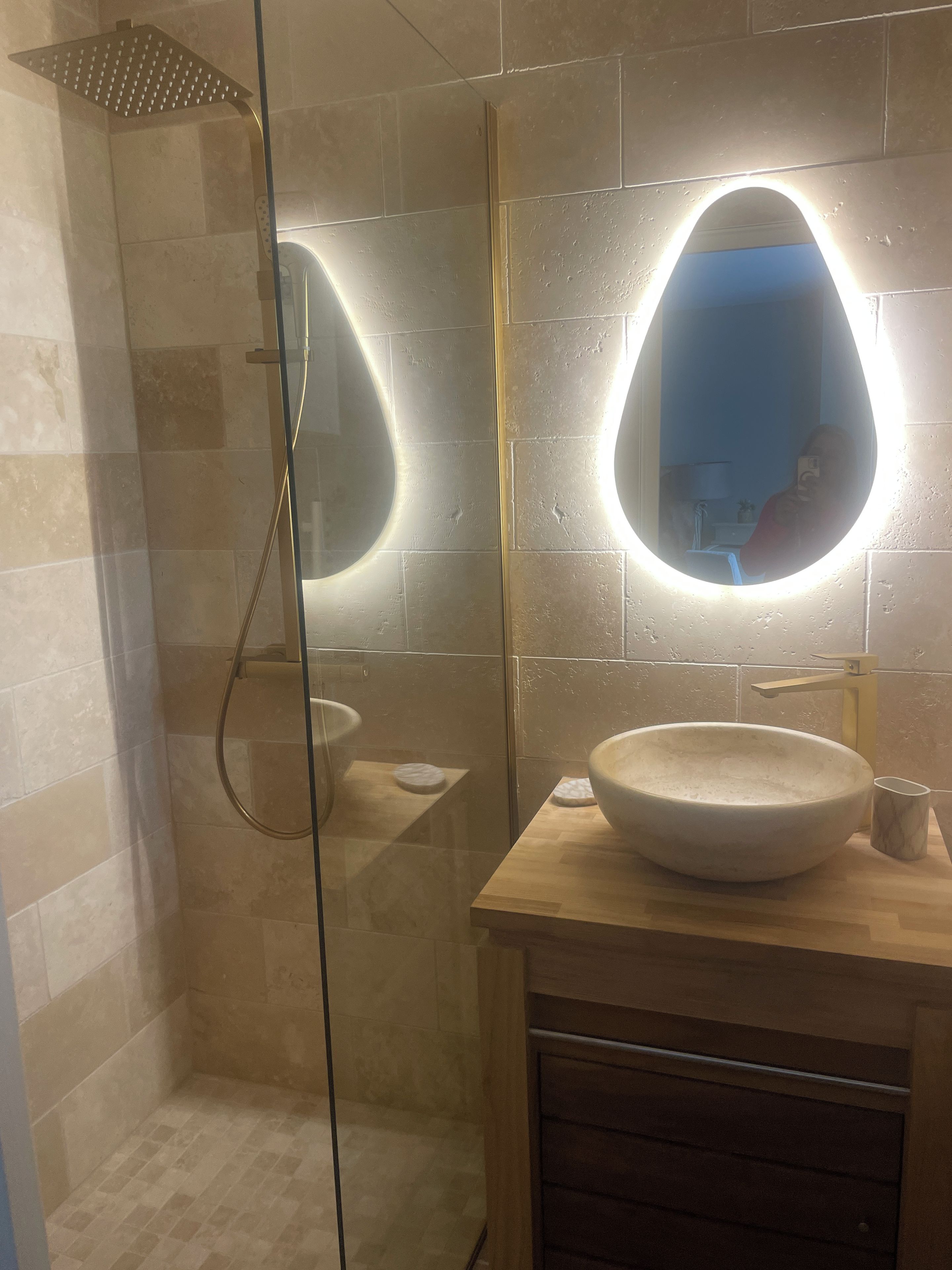 A bathroom with a glass-enclosed shower, a round mirror with a light, and a wooden vanity with a bowl sink.