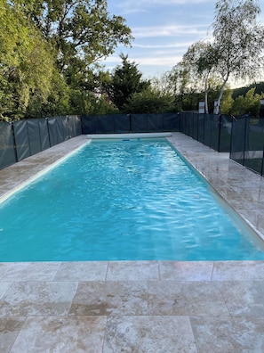 A rectangular swimming pool with a tiled deck, surrounded by a black fence, and trees in the background.